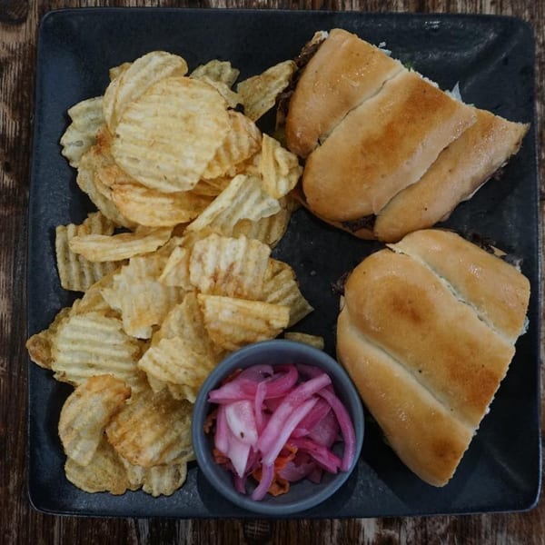 Two sandwiches, a pile of ruffled potato chips, and a small bowl of pickled red onions on a dark plate.