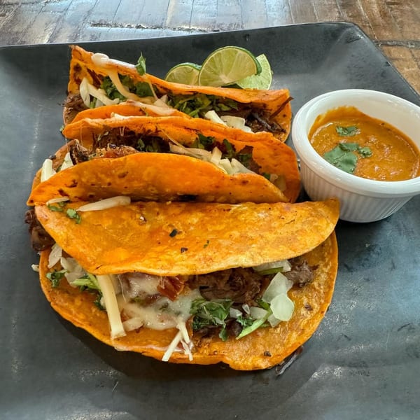 Four birria tacos with lime wedges and a side of consommé on a dark plate.