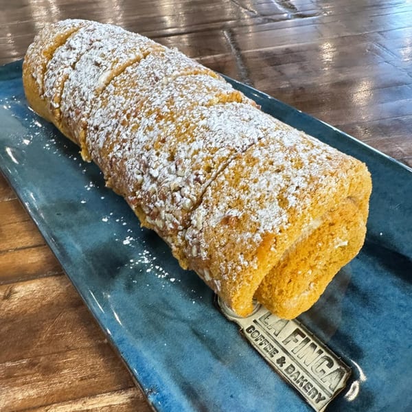 A pumpkin roll cake, dusted with powdered sugar, on a blue rectangular plate from La Finca Coffee & Bakery.